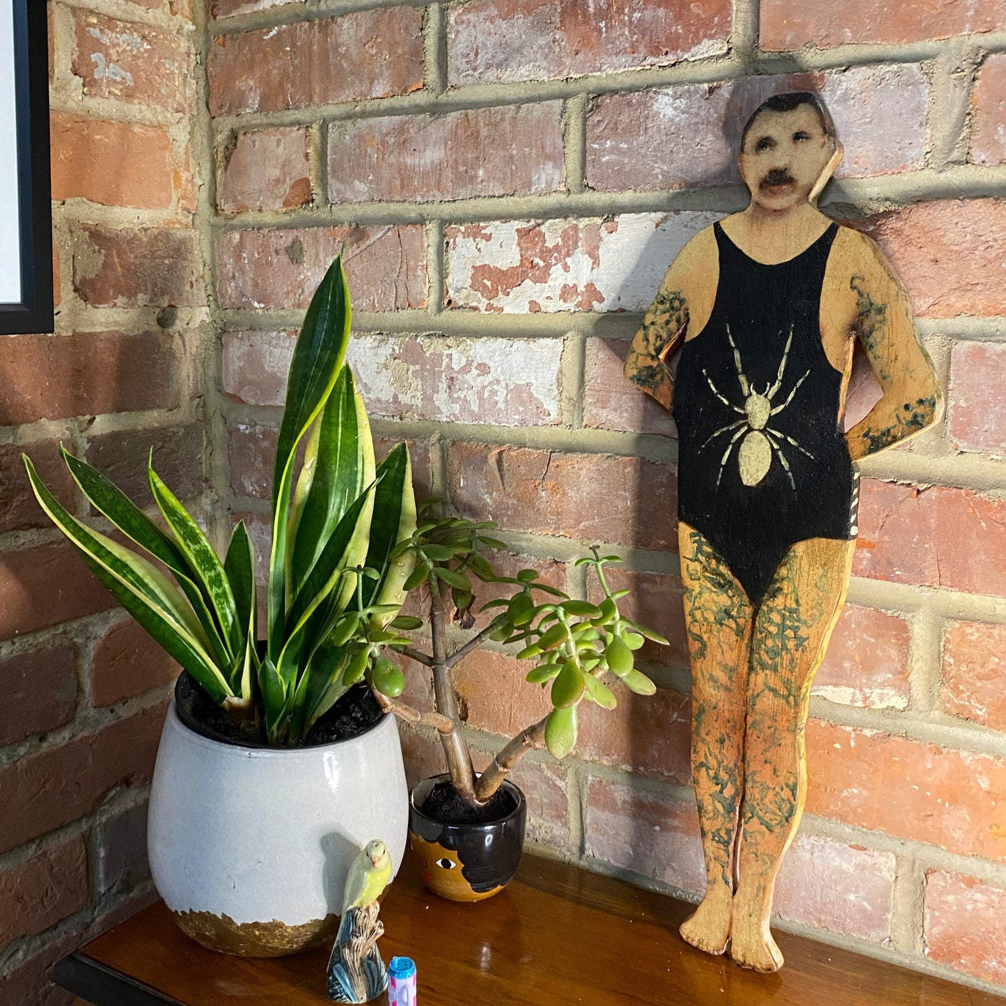 Papillon Man original oil painting on bespoke wood panel, butterfly-themed outsider art with dual-sided narrative, here he's wearing a black swimsuit next to potted plants against a brick wall.