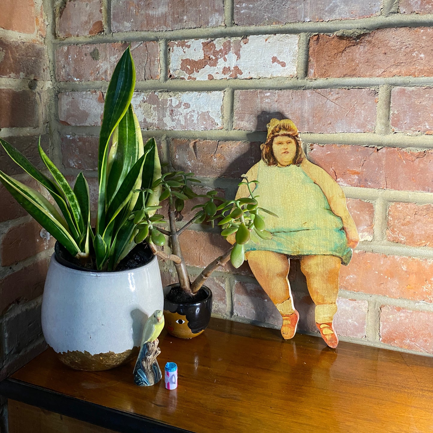 Diane Twinkle Toes original oil painting on wood, circus-inspired outsider art from 1910s-20s era with dual perspectives leaning against a brick wall with plants on a wooden surface.