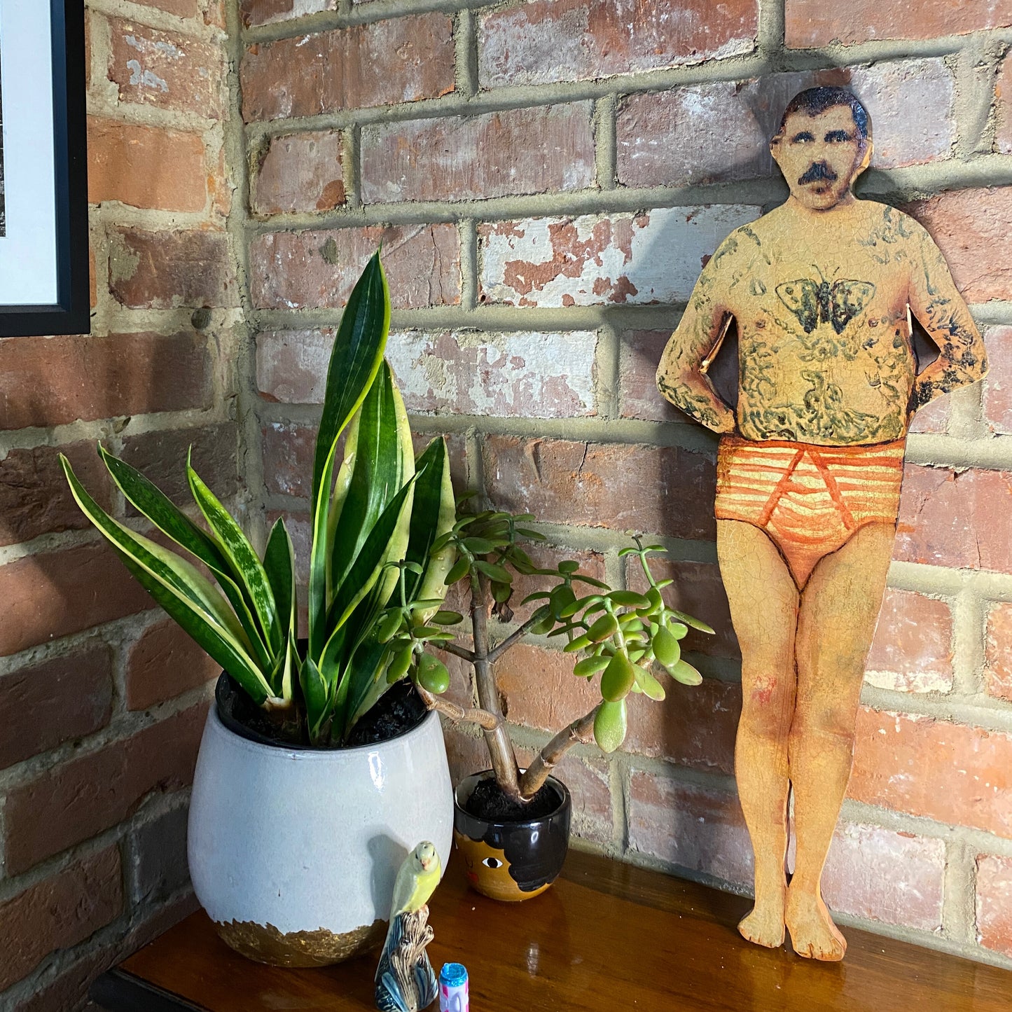 Papillon Man original oil painting on bespoke wood panel, butterfly-themed outsider art with dual-sided narrative, here  he has tattoos and underwear next to potted plants against a brick wall.