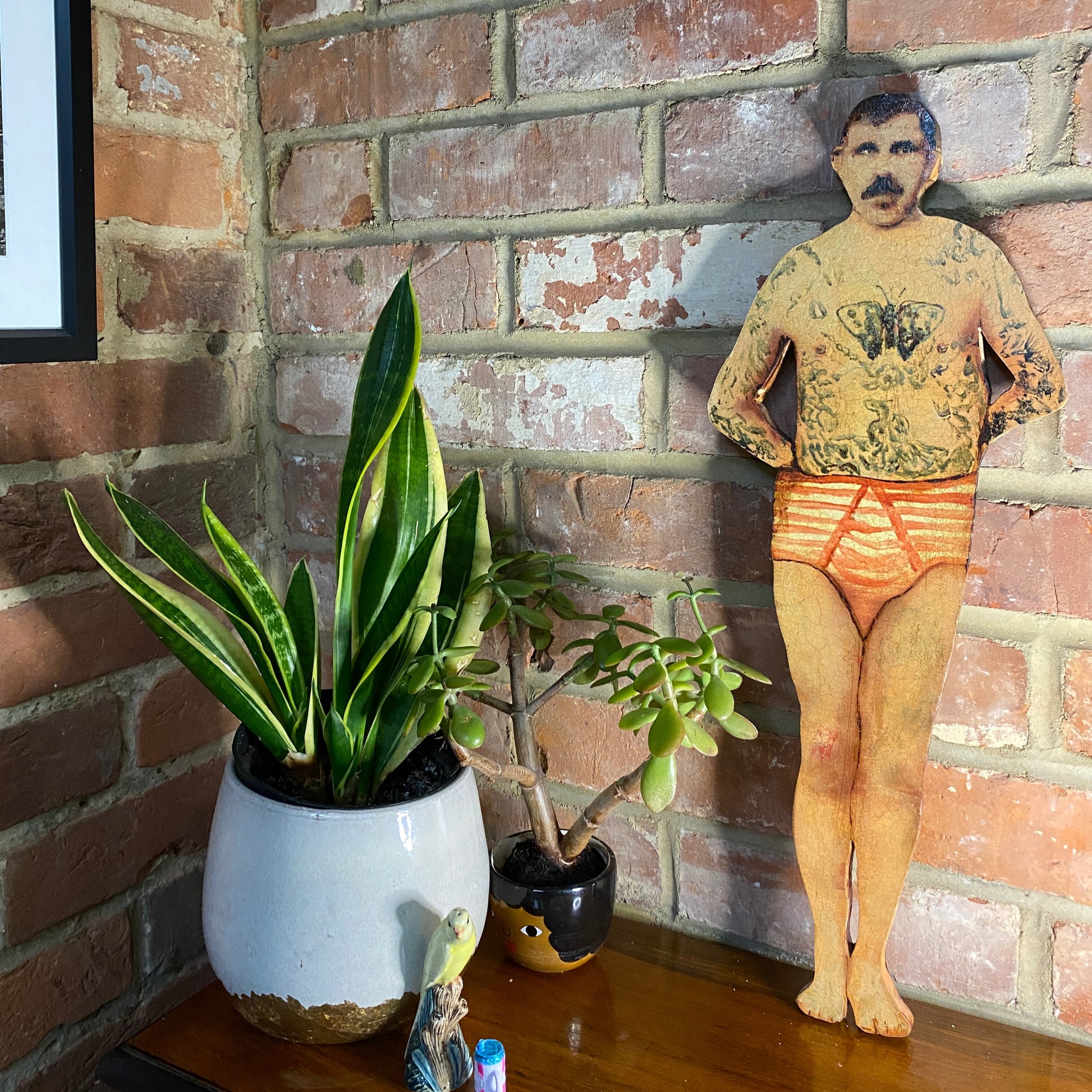 Papillon Man original oil painting on bespoke wood panel, butterfly-themed outsider art with dual-sided narrative, here  he has tattoos and underwear next to potted plants against a brick wall.