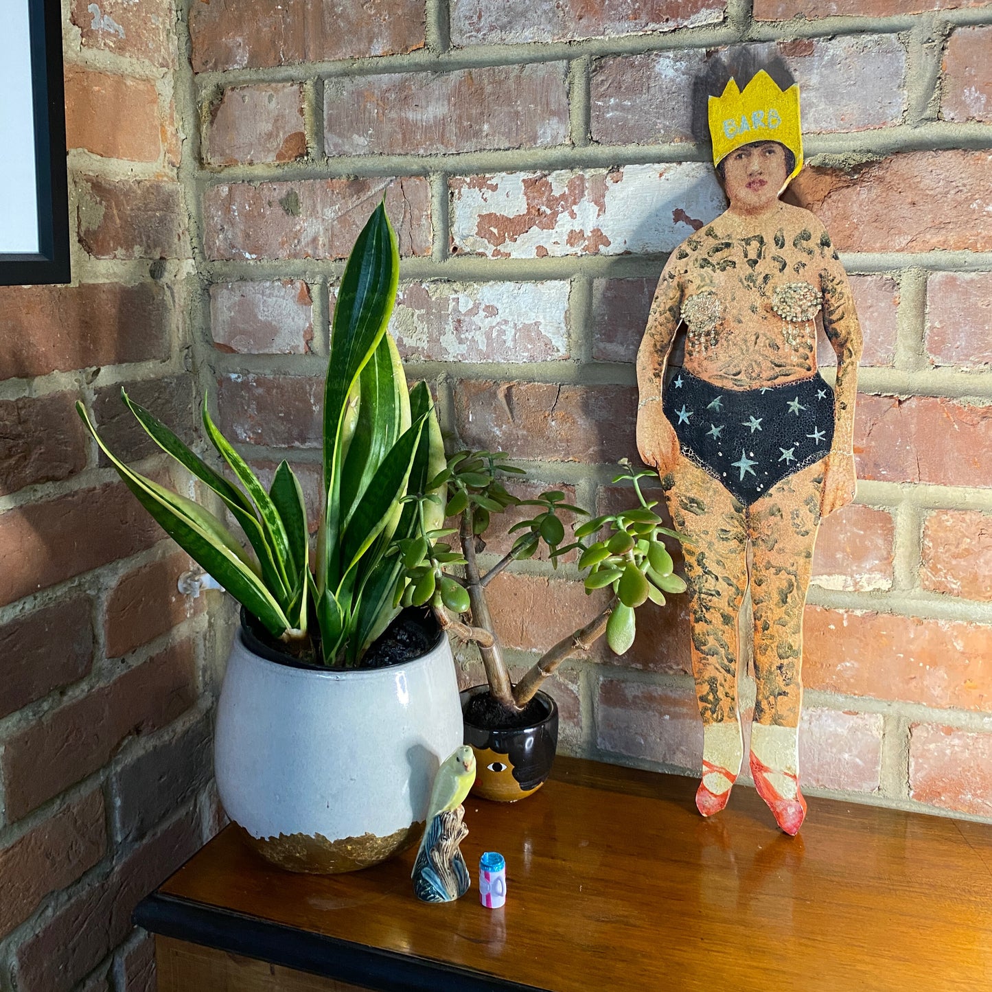 Barb The Queen original oil painting on wood, outsider art featuring regal character with dual-sided storytelling with tattoos and a crown against a brick wall with potted plants on a wooden surface.