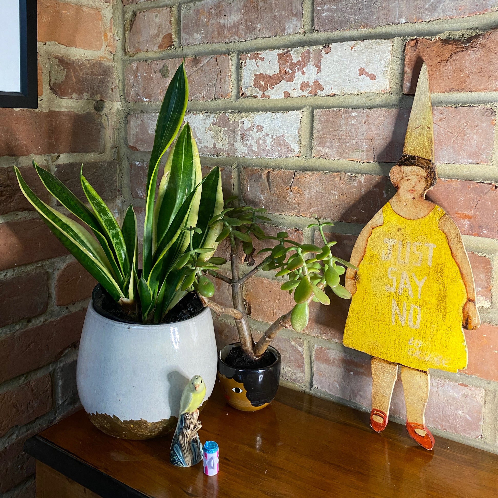 Madge side of Stella, an outsider art piece including a potted plant, small figurines, and a doll against a brick wall.